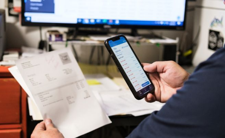 Person scanning an invoice with a phone, with a desk and computer in the background.