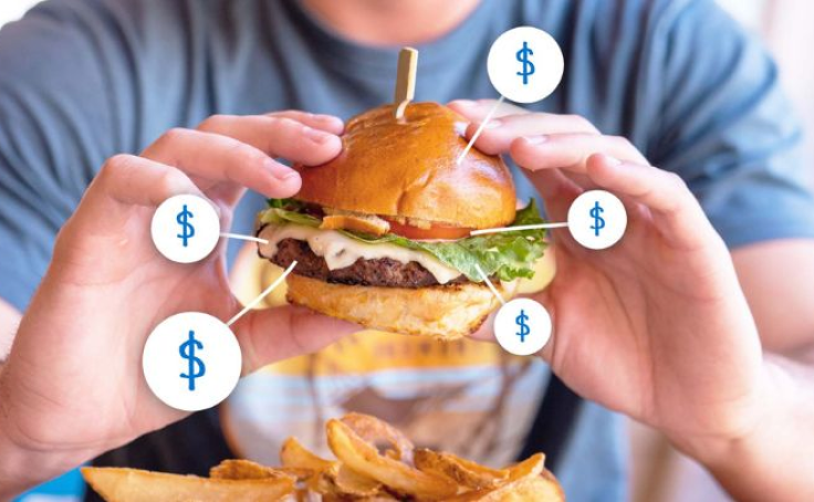 Person holding a burger with dollar signs pointing to ingredients, fries visible in foreground.