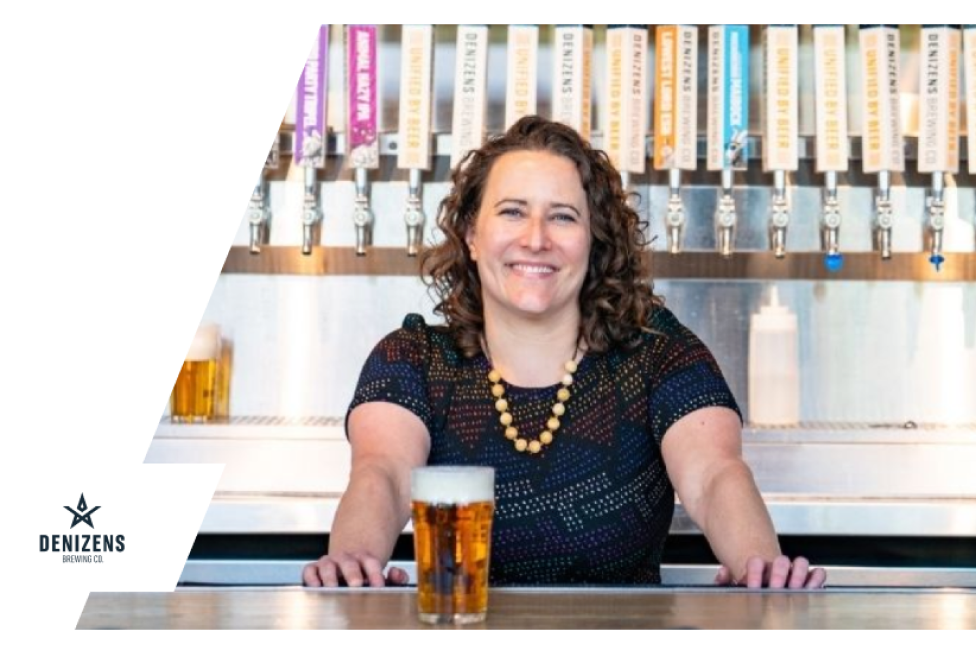 Smiling woman behind a bar with beer taps and a glass of beer, Denizens Brewing Co. logo visible